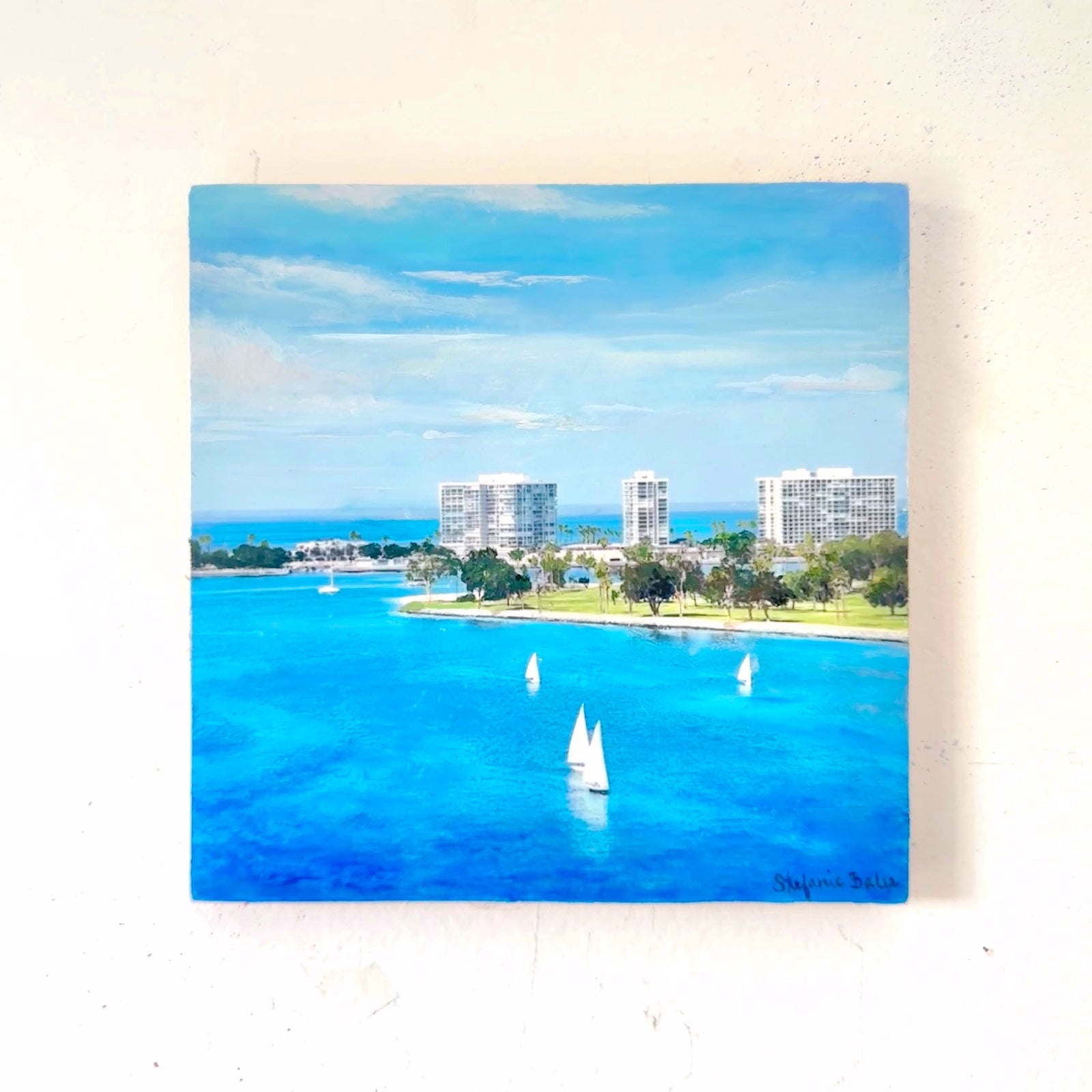 A square painting on wood of a coastal harbor scene  in Coronado, San Diego, California, with bright blue water, sailboats, shoreline trees, and modern buildings under a blue sky, displayed against a white wall.