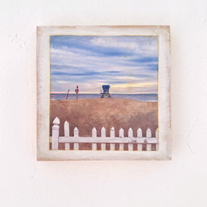 Framed artwork of a  Coronado beach scene  in fron of the Hotel Del with a lifeguard chair, flag, and white picket fence.